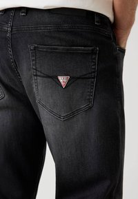 Schwarze Jeans aus Denim verfügen über ein dreieckiges rot-weißes Patch mit dem "Guess"-Logo auf der Gesäßtasche und zeigen ein klassisches Fünf-Taschen-Design.