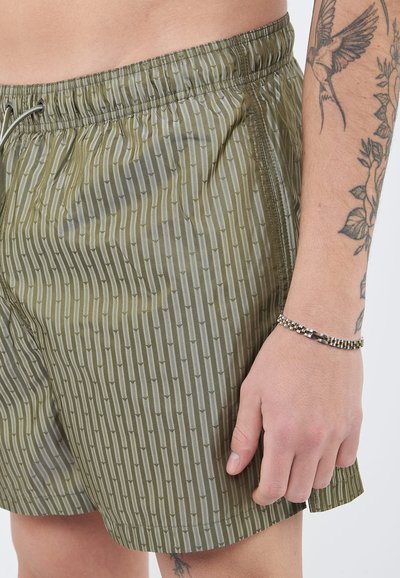 Κοντινό πλάνο ενός ατόμου που φοράει πράσινα ριγέ σορτς με κορδόνι, δείχνοντας τατουάζ στο χέρι και βραχιόλι με χάντρες στον καρπό.