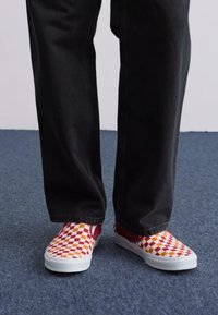 Red and yellow checkerboard slip-on shoes with white rubber soles, worn with black jeans, on a blue carpeted surface.