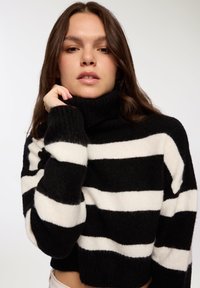 Czarny i biały sweter z golfem w paski, wykonany z miękkiego, puszystego materiału. Posiada długie rękawy oraz skrócony krój z prążkowanym dołem.