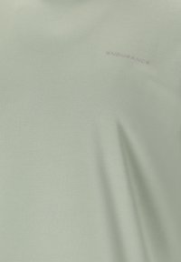 Lichtgroene stof met subtiele tekst "ENDURANCE" gedrukt nabij de rechterbovenhoek, met een gladde textuur en lichte plooien.