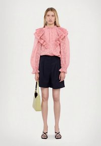 Roosa volangidega pluus pikkade varrukatega, kombineeritud tumesiniste lühikestega. Modell hoiab helerohelist käekotti ja kannab musti rihmikuid sandaale.