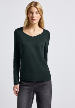 Dunkelgrünes Langarmoberteil mit V-Ausschnitt, glatter Textur, figurbetontem Schnitt und minimalistischem Stil, kombiniert mit grauen Hosen.