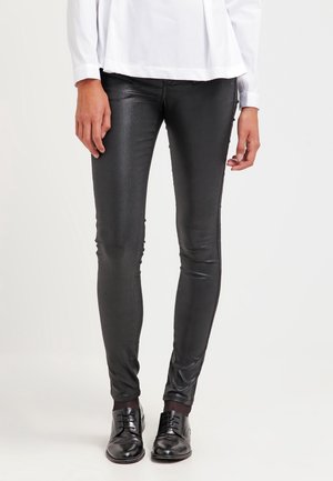 Femme vêtue d'un pantalon skinny noir enduit, de chaussures noires à lacets et d'un chemisier blanc à manches longues, debout devant un fond uni.
