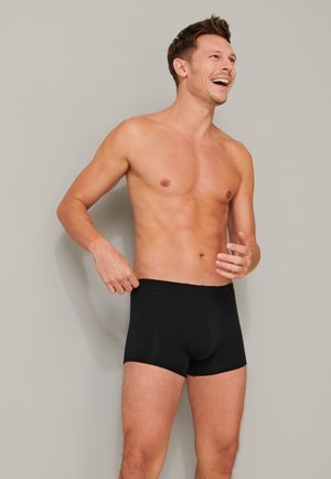 Boxer-briefs noirs en tissu doux, dotés d'une taille sans couture et d'une coupe ajustée, portés par une personne sur un fond neutre.