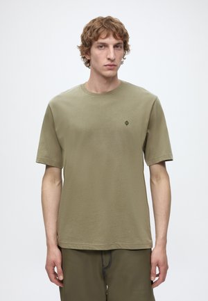 Jeune homme aux cheveux bouclés clairs, portant un t-shirt vert olive uni à manches courtes et un pantalon assorti, debout devant un fond blanc.