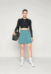 Top corto negro de manga larga, falda deportiva teal con detalles en blanco y zapatillas deportivas blancas. La modelo sostiene una bolsa de gimnasio negra.