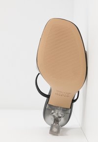 Semelle d'une sandale noire à talon haut avec talon carré transparent, dessous beige texturé et bride chevillère noire, posée sur une surface blanche dans un angle de mur.