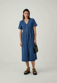 Ženska, oblečena v modro midi obleko s kratkimi rokavi, ki ima gumbe spredaj, drži črno torbico in nosi rjave sandale.