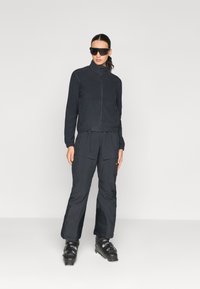 Svart fleecejacka med dragkedja, i kombination med matchande svarta cargobyxor och svarta skidstövlar. Solglasögon fulländar outfiten.