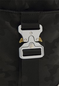 Borsa in nylon camouflage nera con una fibbia metallica in argento e oro, caratterizzata da una superficie testurizzata e una tracolla in tessuto nero.