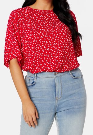 Femme portant un chemisier rouge à manches courtes avec de petits motifs floraux blancs et un jean taille haute bleu clair, main reposant à côté.