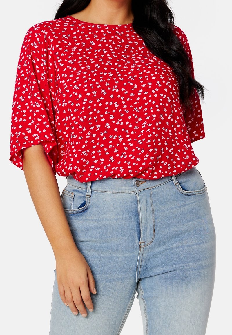 Femme portant un chemisier rouge à manches courtes avec de petits motifs floraux blancs et un jean taille haute bleu clair, main reposant à côté.