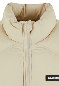 Chaqueta acolchada beige con collar alto, cierre de cremallera completa y una etiqueta de marca negra en la parte delantera. Textura suave y diseño acolchado.