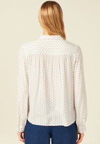 Blusa di chiffon chiara con motivo a pois blu, maniche lunghe, schiena plissettata e collo classico. Il tessuto è liscio, leggero e leggermente trasparente.