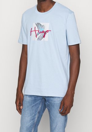 Mężczyzna w jasnoniebieskiej koszulce z niebieską papugą i czerwoną, kursywą napisaną nazwą "Hugo" z przodu, w komplecie z niebieskimi dżinsami, na tle jednolitego tła.