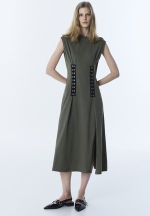 Joven mujer con vestido midi sin mangas de color verde oliva con detalles de cierre de gancho y ojo negros y zapatos slingback negros de punta afilada.