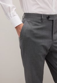 Pantaloni grigi su misura in tessuto strutturato, con tasca discreta e chiusura con bottone, abbinati a una camicia bianca a maniche lunghe.