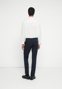 Fehér hosszú ujjú ing és tengerészkék nadrág. A nadrágokon hátul gombbal záródó zsebek találhatók. A megjelenést fekete cipők egészítik ki.