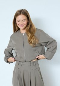 Graue, taillierte Jacke mit silbernem Frontreißverschluss, elastischen Ärmeln und seitlichen Taschen, kombiniert mit passenden plissierten Hosen.