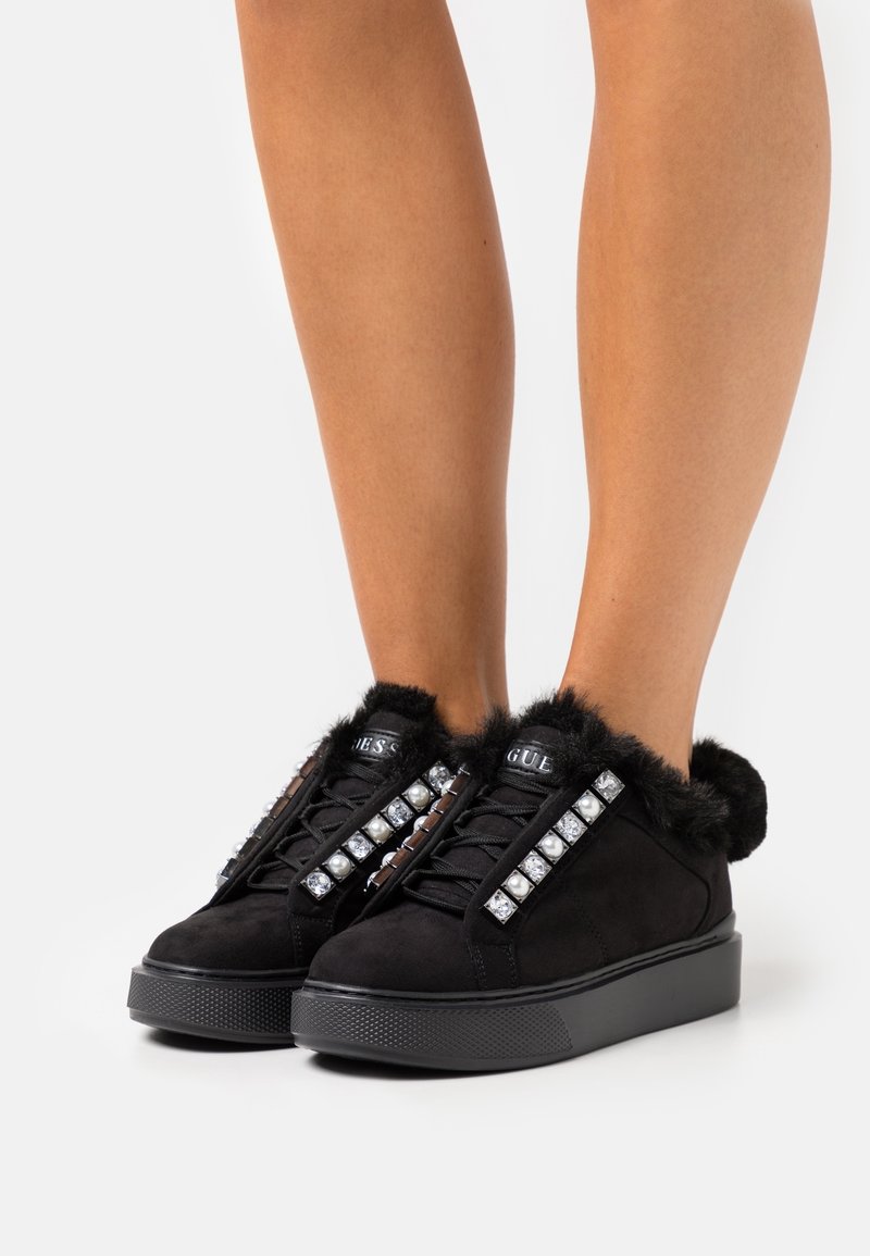 Pieds chaussés de baskets à semelle compensée noires avec garniture en fausse fourrure et ornements décoratifs en perles et pierres précieuses sur fond blanc.