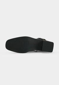 Semelle de chaussure noire avec un talon carré et des détails antidérapants texturés, vue par en dessous sur un fond blanc uni.