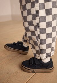 Melni auduma apavi ar divām Velcro siksnām, kopā ar pelēkiem un baltiem rūtainiem biksēm. Apaviem ir brūngana gumijas zole.