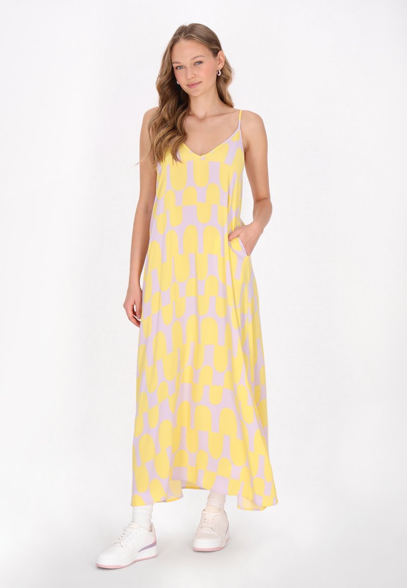 Langes, gelbes Kleid mit lila abstraktem Muster, leichtes Material, Spaghettiträger, seitliche Taschen. Kombiniert mit weißen Sneakern.