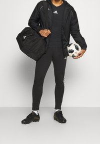 Schwarze, wasserdichte Jacke, schwarzes, eng anliegendes Shirt, schwarze Leggings mit weißen Streifen, schwarze Fußballschuhe, hält einen Fu�ßball und eine Sporttasche.