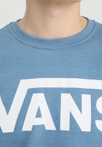 Světlě modrá mikina s kulatým výstřihem, na níž je velké bílé logo "VANS". Tkanina vypadá měkce, s žebrovaným detailem na výstřihu.