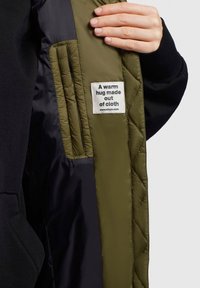 Innenfutter der gesteppten Jacke in Olivgrün mit schwarzen Akzenten. Mit einem Etikett, auf dem steht: "Eine warme Umarmung aus Stoff." Glatte Textur.