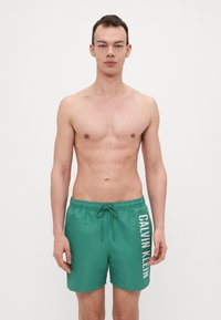 Grønne svømmedrakter med elastisk talje og snøring, med stort hvitt "CALVIN KLEIN"-tekst på venstre side. Glatt stoff.