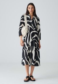 Schwarz-weiß gemustertes Kleid mit einem gestuften Design, Kragen und langen Ärmeln. Kombiniert mit einer weißen Tasche und schwarzen Sandalen.