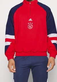 Chaqueta cortavientos roja y marina con cuello con cremallera, puños elásticos y dobladillo. Presenta detalles de rayas en blanco y azul y un logo bordado.