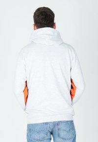 Muž oblečený v světlém šedém svetru s oranžovými akcenty na rukávech a modrých džínách, stojící s turned zády k fotoaparátu.