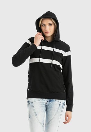 Frau trägt einen schwarzen Kapuzenpullover mit zwei weißen horizontalen Streifen und hellblauen Jeans mit Acid-Waschung, hält eine Kapuzenkordel fest.