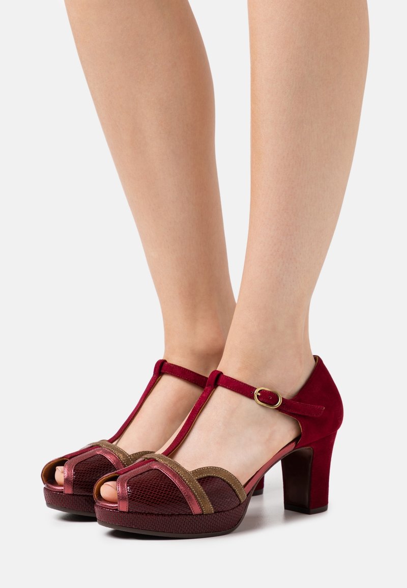 Pieds portant des sandales à talons hauts à bride en T rouges avec des accents marron et rose métallique, debout sur un fond blanc uni.