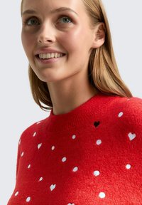 Roter Pullover mit strukturiertem Strick, der weiße und graue Herzmuster aufweist. Hoher Kragen, eng anliegender Schnitt und weiche, gleichmäßige Oberflächenstruktur.