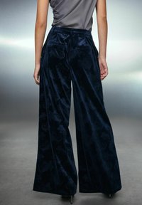 Woman wearing high-waisted, wide-leg dark blue velvet pants with subtle floral patterns, paired with a gray sleeveless top.