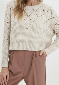 Světlý béžový pletený svetr s geometrickými vzory a žebrovaným lemem. Kombinován s volnými, měkkými hnědými kalhotami, vytvářející texturovaný vzhled.