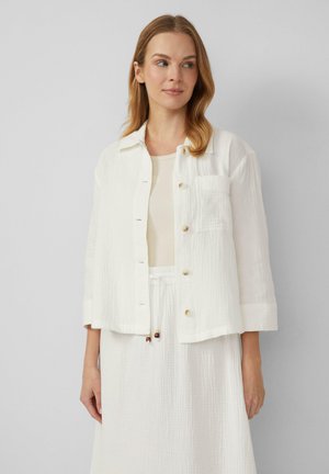 White cropped jacket made of textured fabric, featuring a collar, button closure, and a front pocket. Worn over a matching light top.