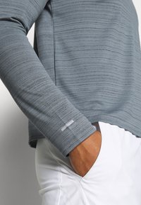 Graues langärmeliges Hemd mit einem strukturierten Design, ausgestattet mit einem dezenten weißen Etikett am Ärmel und einem lockeren Schnitt, kombiniert mit weißen Hosen.