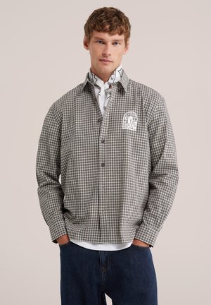 Jeune homme aux cheveux bouclés portant une chemise à carreaux boutonnée avec le texte "BLUE RIDGE" et un foulard blanc à motif autour du cou.