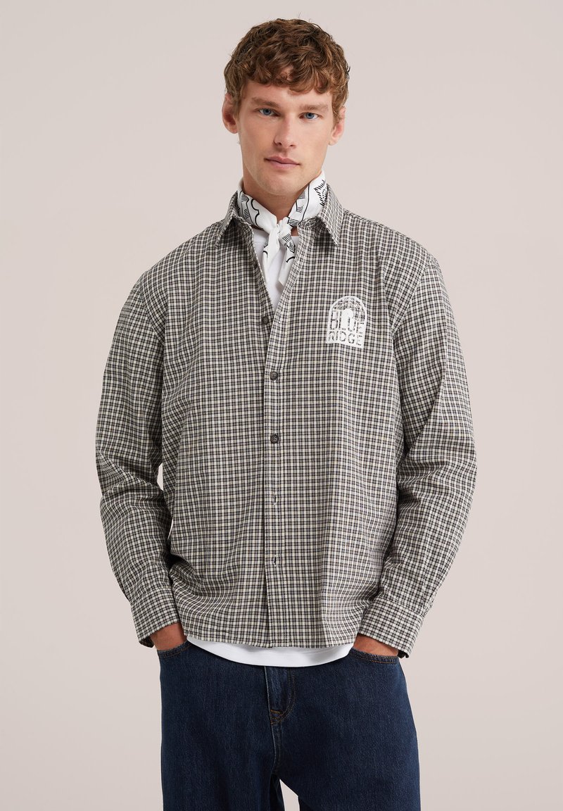 Jeune homme aux cheveux bouclés portant une chemise à carreaux boutonnée avec le texte "BLUE RIDGE" et un foulard blanc à motif autour du cou.