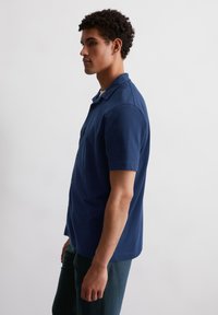 Jonge man in profiel, gekleed in een textuurnavyblauwe, kort aangesloten overhemd met knopen en donkere broeken, tegen een plain witte achtergrond.
