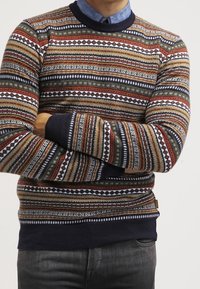 Pull en maille multicolore avec des motifs à rayures horizontales en rouge, vert et bleu. Poignets et ourlet côtelés bleu marine, tissu texturé.