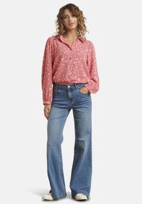 Chemise rose à manches longues avec un motif en forme de cœur, associée à un jean évasé bleu clair. Le mannequin se tient avec les mains dans les poches, portant des baskets blanches.