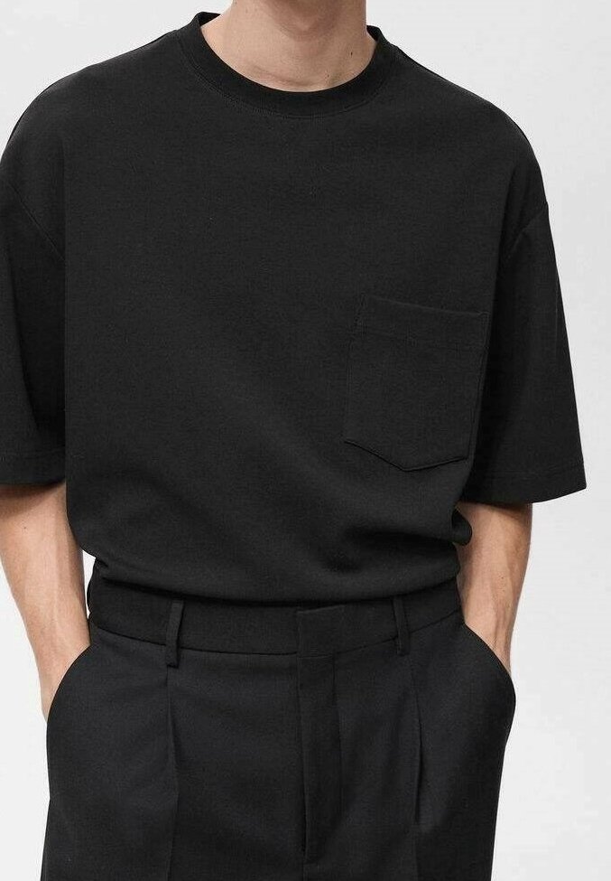Homme portant un t-shirt noir uni à manches courtes avec poche poitrine et pantalon noir taille haute, mains dans les poches, sur fond clair.