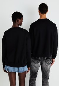 Svart hettegenser med senket skuldre, brukt av to personer. Den ene er kombinert med et denimskjørt, den andre med slitte grå jeans.