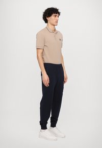 Beige polo skjorte med krage, logo og korte ermer, samt marineblå joggebukser og hvite sneakers. Enkel, funksjonell design.